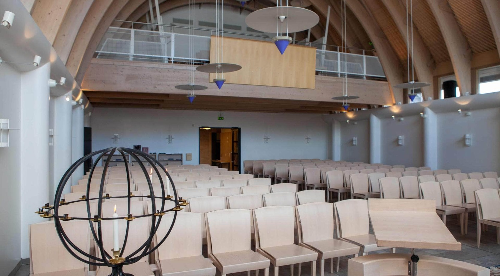 Spacious hall with curved wooden vaulted ceiling, rows of light-wood chairs, mezzanine balcony and modern pendant lights with blue cone accents.