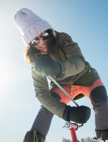 Smiling guest in knit hat and sunglasses drilling an ice hole with an auger on a sunny winter day