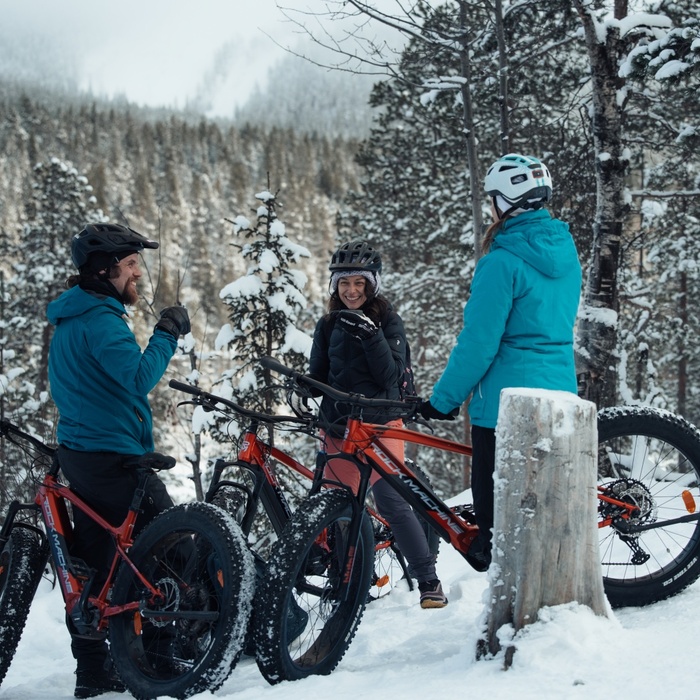 Guests on a guided fat‑bike tour in a snowy mountain forest, wearing helmets and winter jackets, laughing beside red bikes