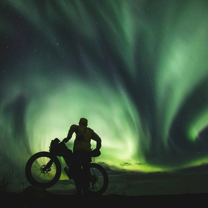 Cyclist silhouette with a fat‑tire bike beneath swirling green northern lights - remote aurora-viewing adventure for guided tours.