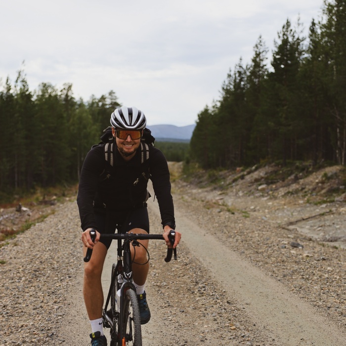 Cyclist riding a gravel forest road, wearing helmet, sunglasses and backpack with distant hills under a cloudy sky