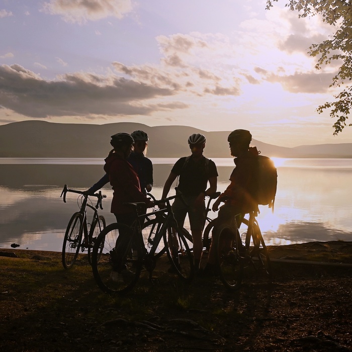 Cyclists silhouetted at a tranquil lakeshore at sunset, mountain views and leafy shore — ideal for hotel bike tours and lakeside excursions.