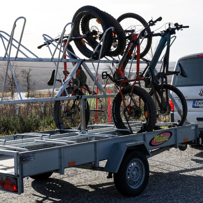 Silver van with a trailer-mounted bike rack carrying several fat‑tire mountain bikes on a gravel roadside with distant hills.