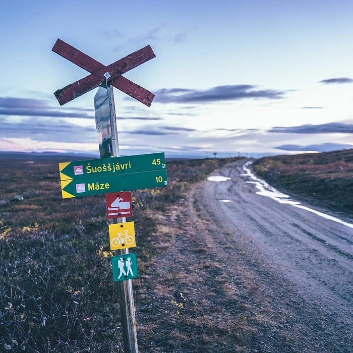 Trail signpost with hiking, cycling and skiing icons and distances to Šuoššjávri (45 km) and Máze (10 km) by a remote gravel road at dusk