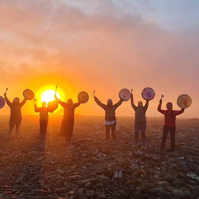 Guests on a rocky plateau hold hand drums aloft during a vibrant sunrise drumming ceremony
