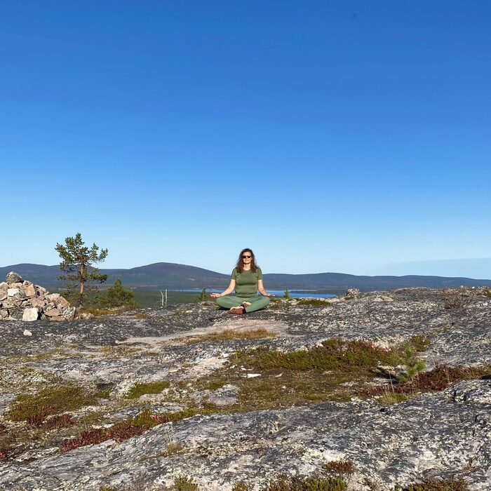 Woman meditating in green yoga clothes on rocky hilltop terrace with lake and distant forested mountains under clear blue sky