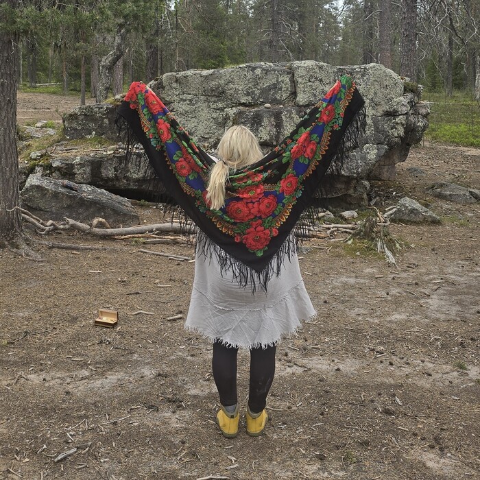 Guest in forest holding colorful floral shawl before lichen-covered rock, hiking trail ambiance