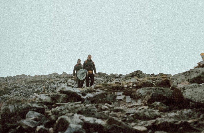 Two hikers carrying round boards across a rocky, remote hillside under a pale overcast sky