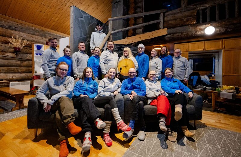 Lodge team gathered in a cozy log-cabin lounge with wooden beams, stone stair, matching fleece jackets, wool socks and communal sofa.