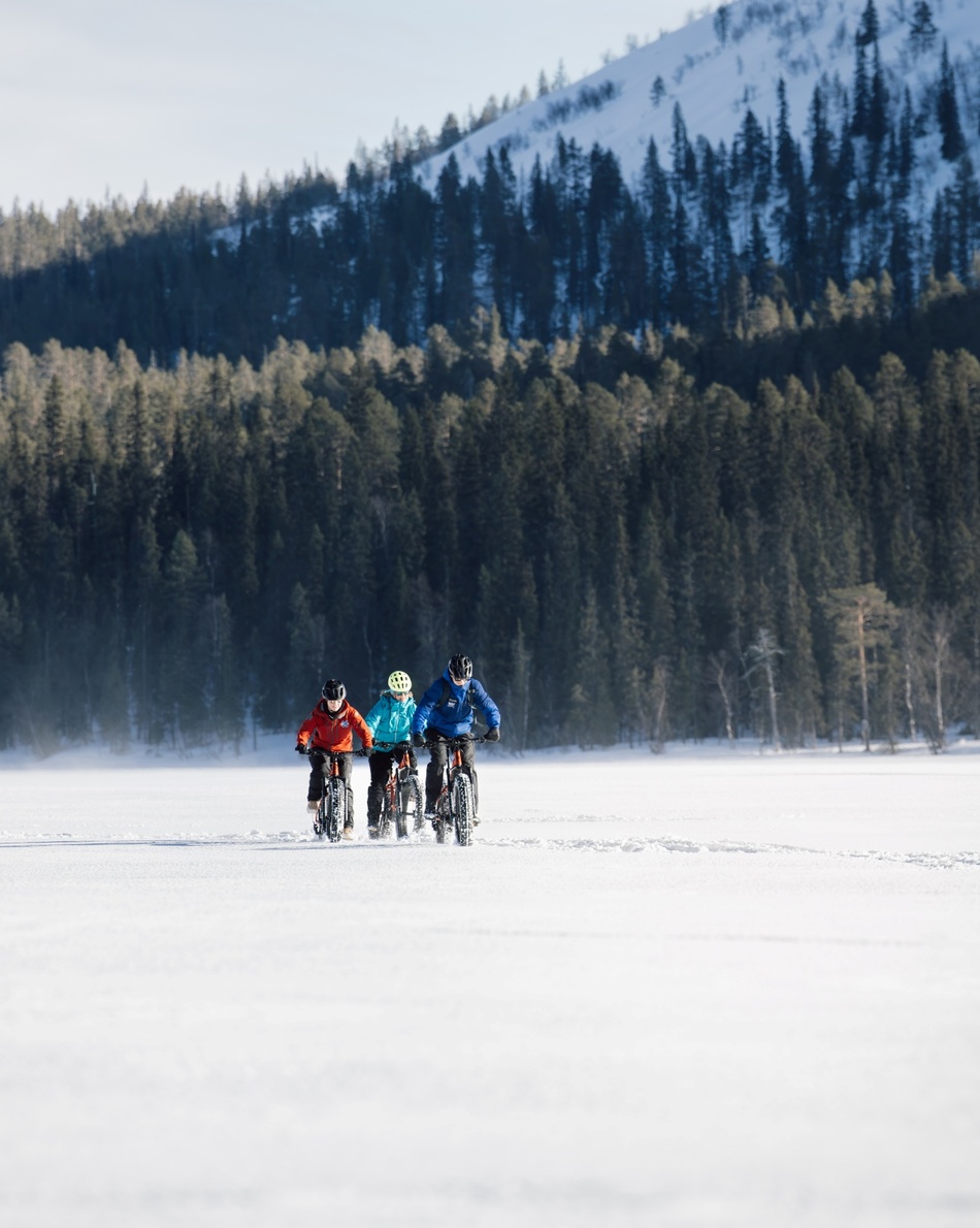 Guided fat-bike ride across a snowy frozen lake, three cyclists in colorful winter jackets with forest and mountain backdrop