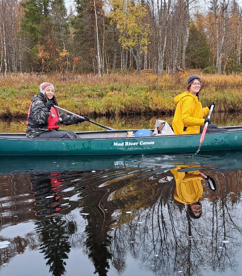 Two hotel guests paddling a green canoe on a calm autumn river, wearing jackets and life vests, birch forest reflections.