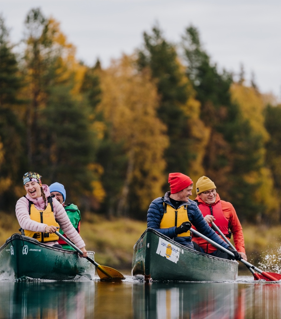 Guests on a guided canoe trip paddle across a calm lake amid golden autumn forest, smiling and wearing life jackets.