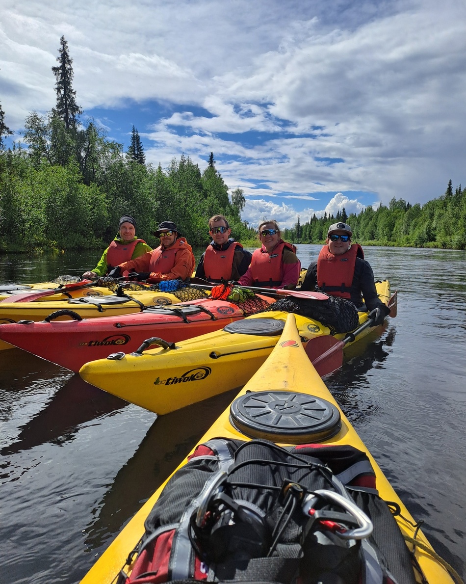 Five paddlers in yellow and red kayaks wearing life jackets on a calm river, surrounded by green forest under a blue sky.