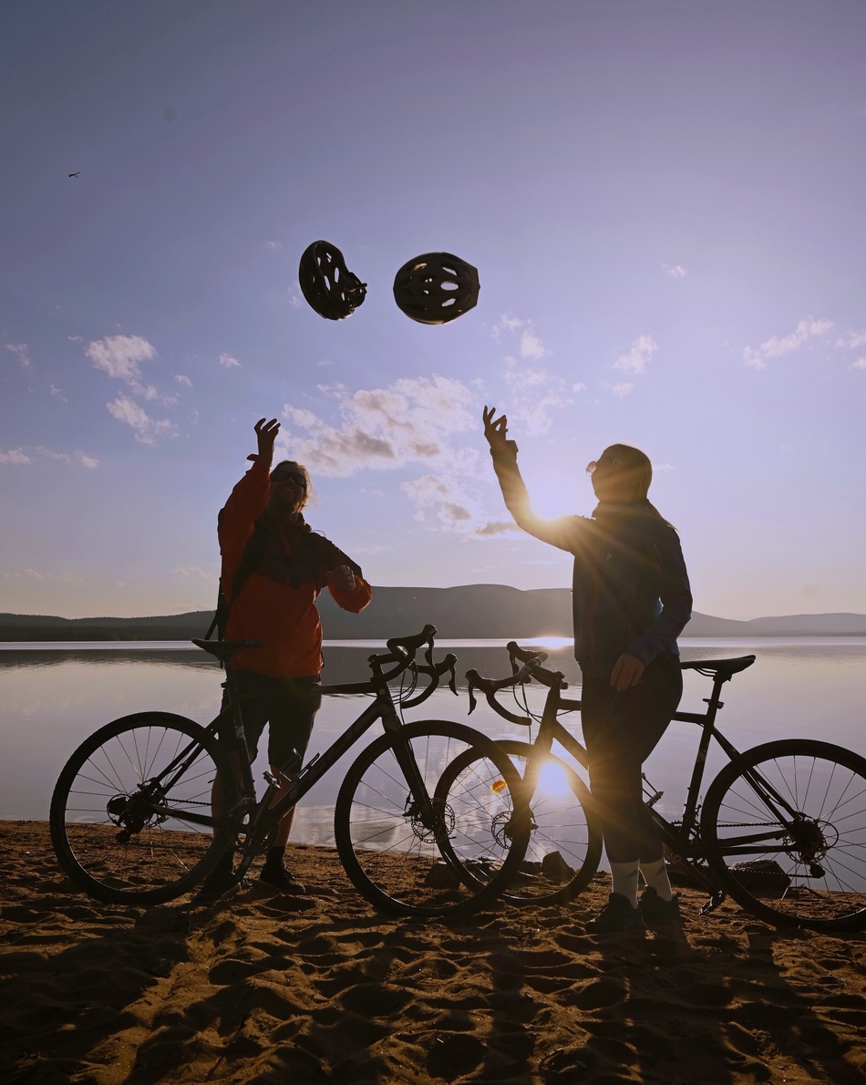 Two cyclists toss helmets by their bikes on a lakeside beach at sunset, with calm water and distant mountains.
