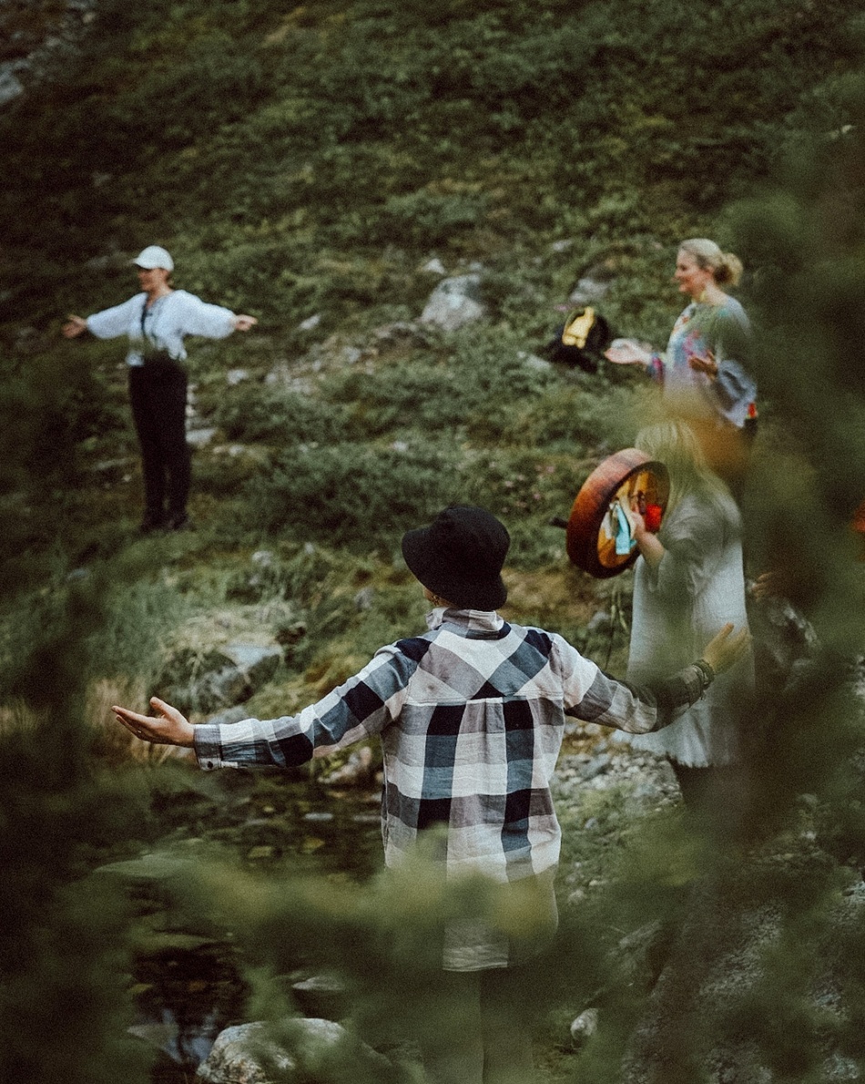 Guests at a mountain retreat on a grassy slope, arms outstretched during an outdoor drumming and wellness circle