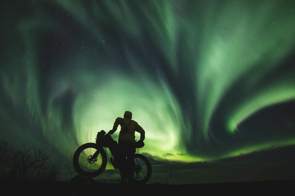 Cyclist silhouette with a fat‑tire bike beneath swirling green northern lights - remote aurora-viewing adventure for guided tours.