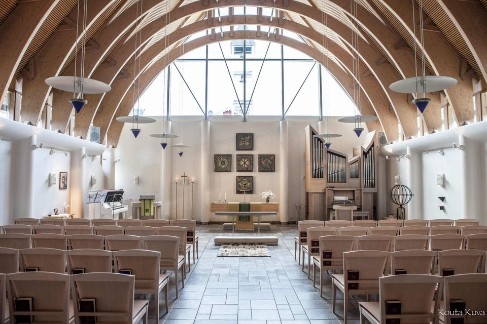 Bright chapel with soaring wooden arches, large arched window, pipe organ, simple altar and rows of pale wooden chairs.