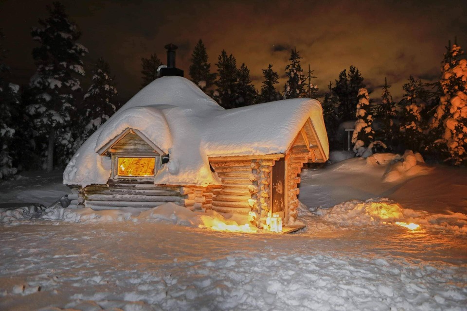 Snow-covered log cabin with thick roof drift, warm glowing window and lanterns, secluded forest setting at night