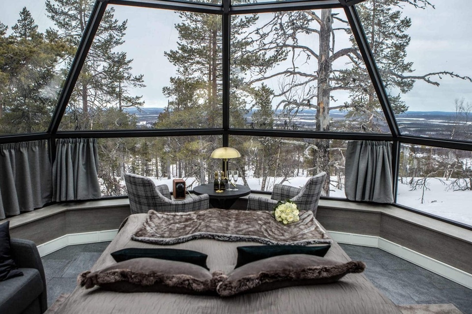Panoramic glass suite with king bed, fur throw, two plaid chairs and table set with champagne, snowy pine forest view
