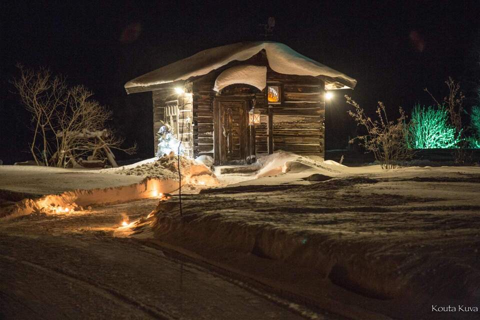 Snow-covered log cabin at night, warmed by lanterns and a candle-lit path, cozy wooden entrance and frosted shrubs