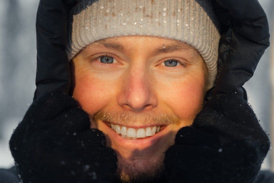 Smiling person in black hood and knit beanie, gloved hands framing face with snowflakes on hat and rosy cheeks