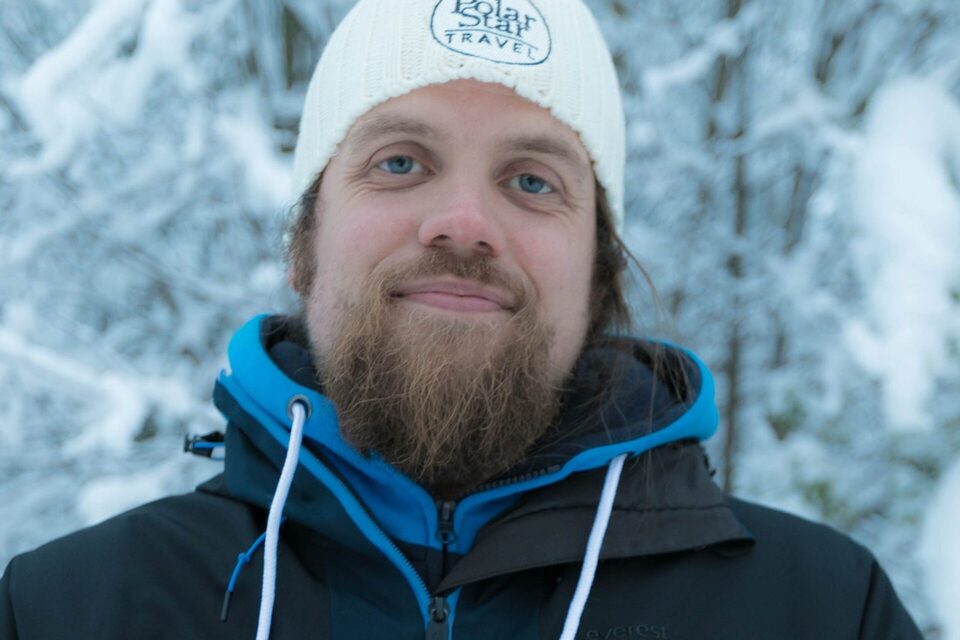 Smiling bearded Polar Star Travel guide in a branded white beanie and blue winter jacket amid a snow-covered forest