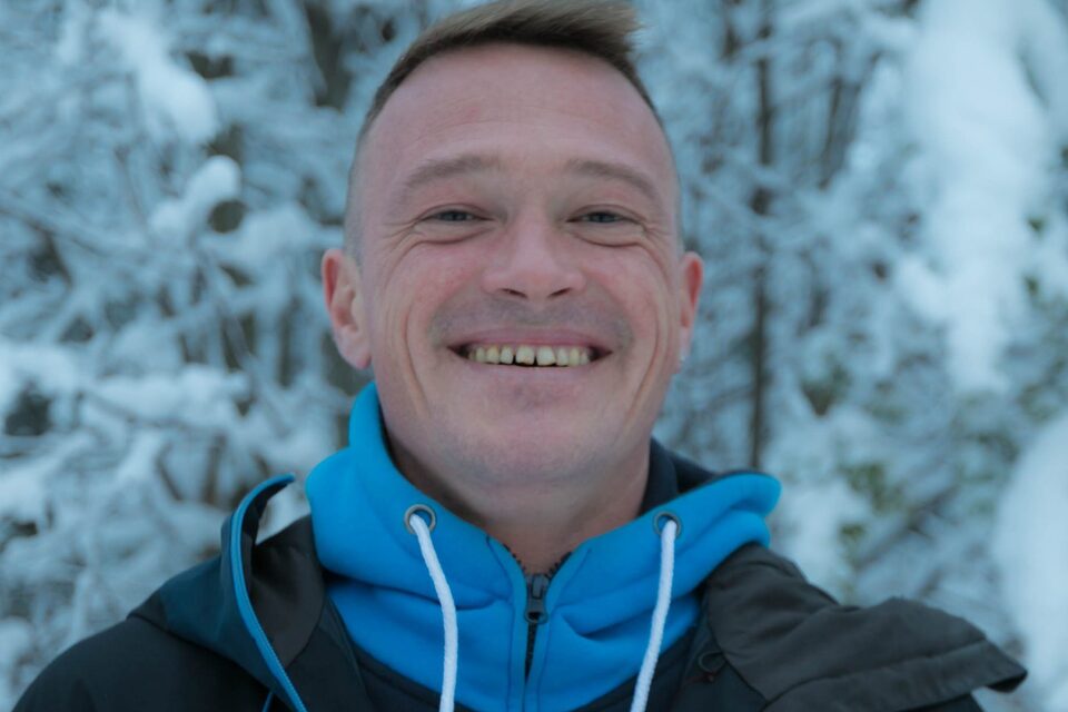 Smiling man in blue hoodie and winter jacket, close-up selfie with snow-covered trees in background