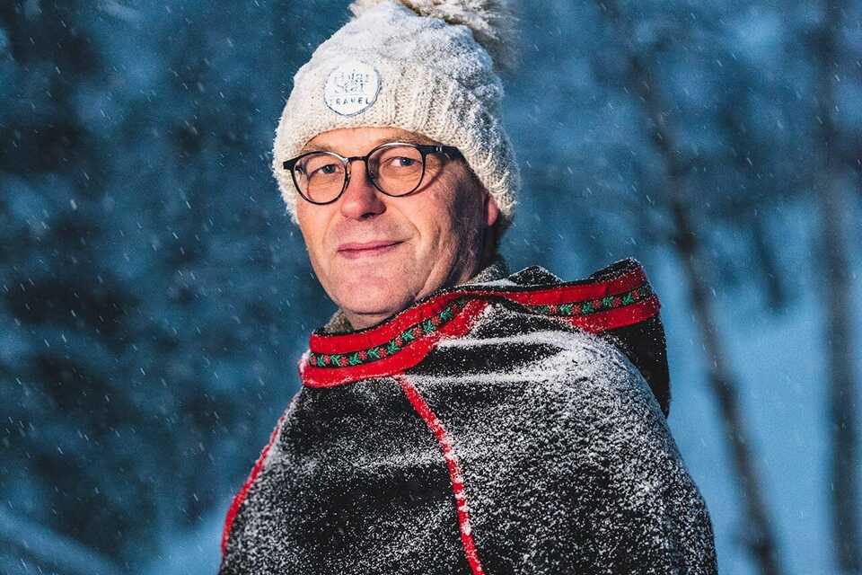 Local winter guide in falling snow, wearing a traditional wool cloak with red trim and a snow-dusted knit pompom hat