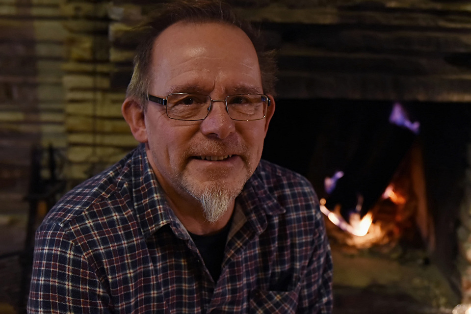 Guest in glasses and plaid shirt smiling by a glowing stone fireplace in a cozy hotel lounge