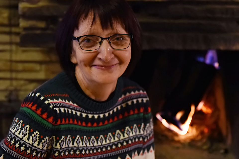 Smiling guest in a patterned winter sweater beside a stone fireplace, warm firelight and cozy lodge ambiance.