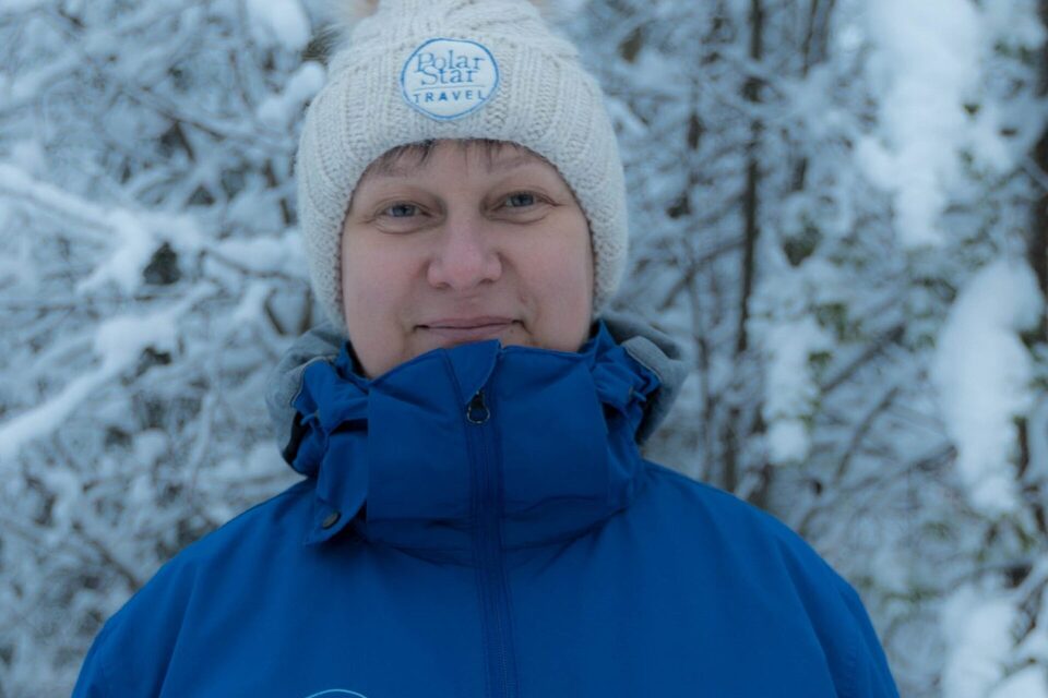 Smiling person wearing a white knit hat with 'Polar Star Travel' badge and blue winter jacket in a snow-covered forest