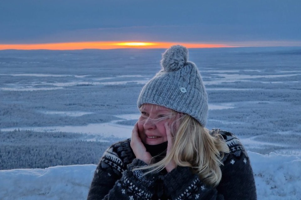 Guest in a gray pom‑pom knit hat and patterned sweater smiling while watching sunset over a vast snowy forest plain