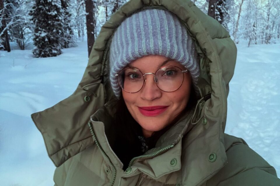 Smiling guest wearing glasses, green puffer coat and knit beanie on a snowy forest walk at dusk