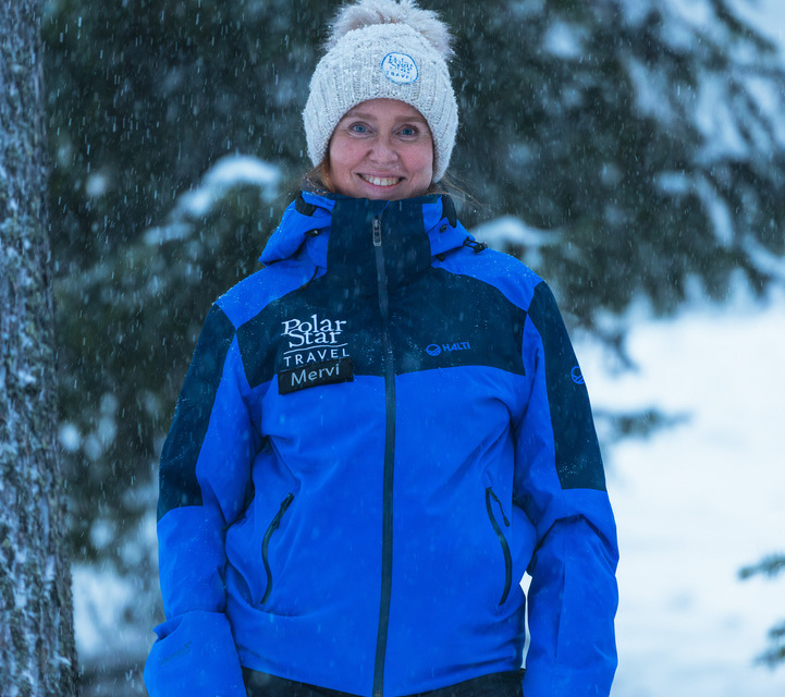 Smiling winter tour guide in blue Polar Star Travel jacket and knit beanie standing in falling snow among snow-covered pines