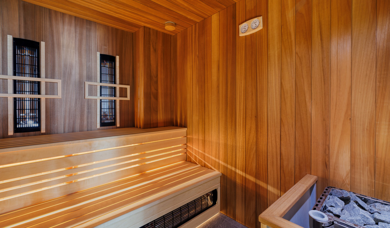 Private wooden sauna with tiered bench seating, warm wood paneling, infrared panels, heater with stones and wall thermometer