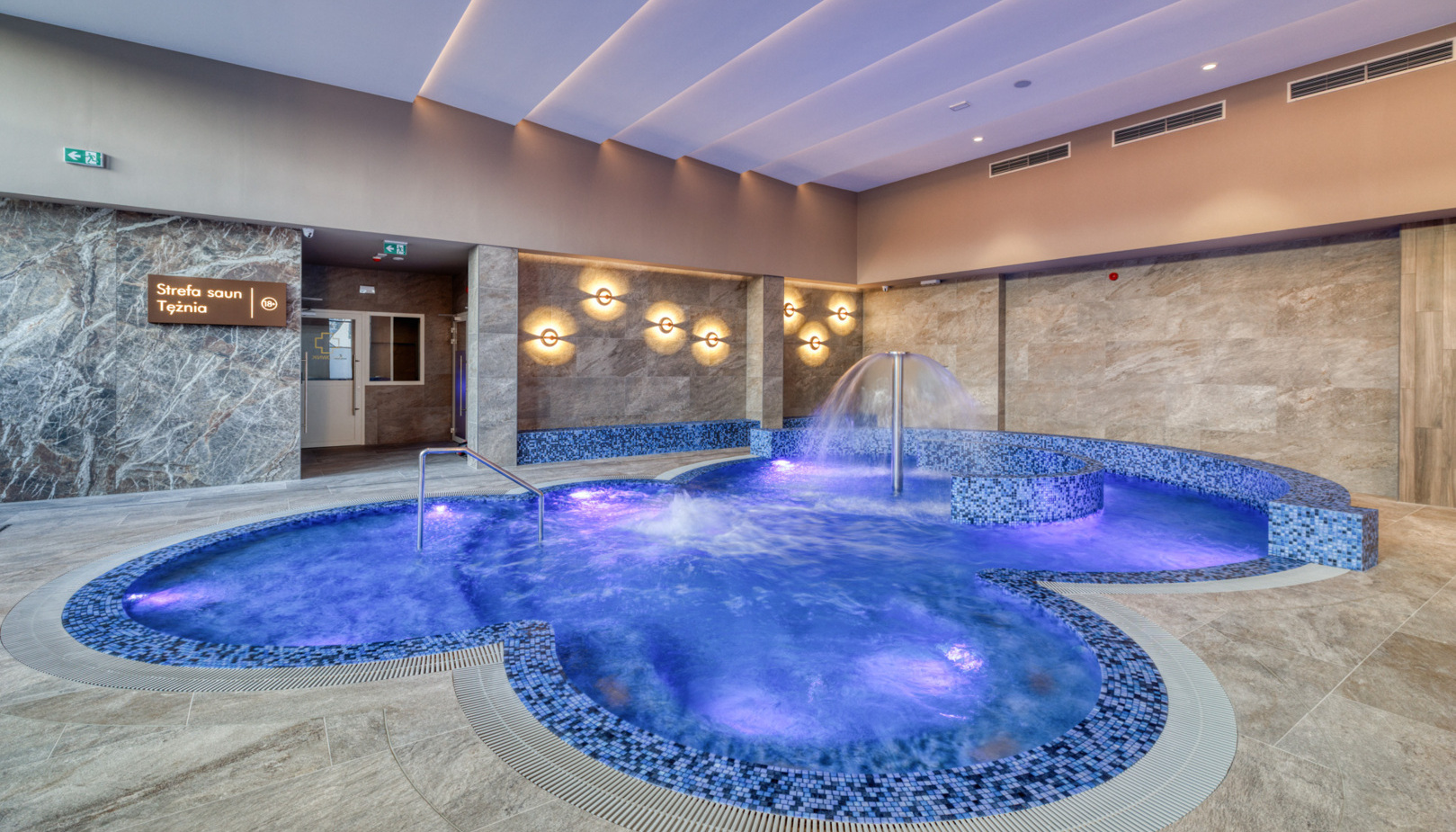 Indoor spa pool with mosaic tile trim, cascading water feature and purple LED lighting; marble walls and entrance sign to sauna area.
