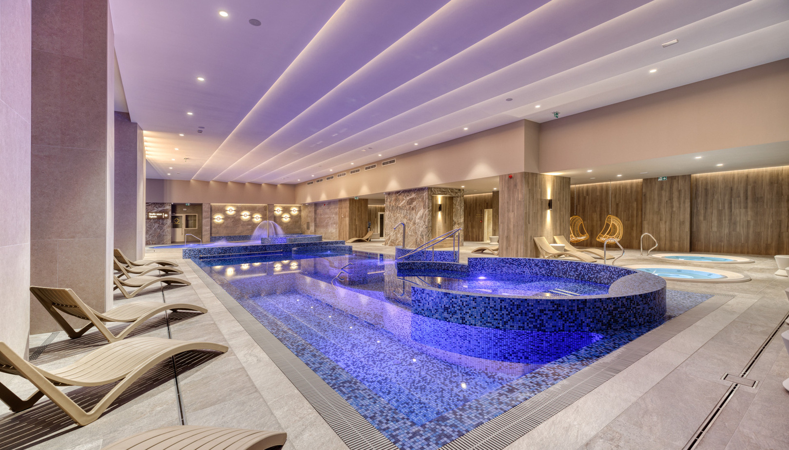 Luxurious indoor spa with mosaic-tiled pool and raised whirlpool, ambient purple lighting, wooden walls and reclining loungers.