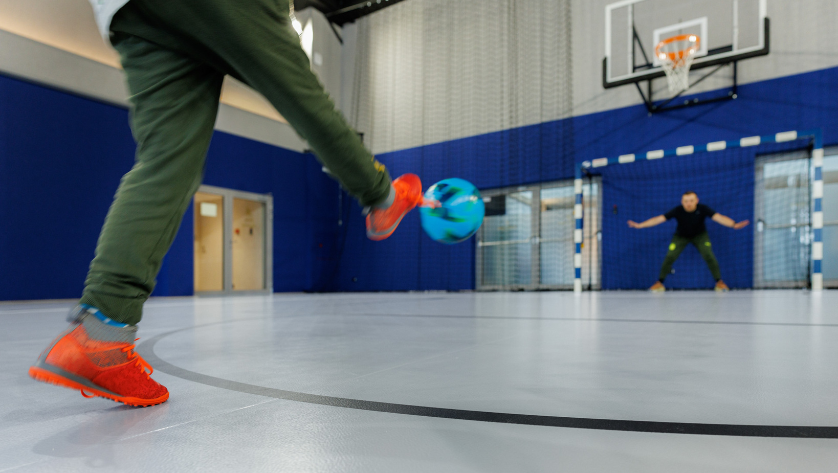 Indoor multipurpose court: guest in orange sneakers kicks a colorful ball toward a goalkeeper under a basketball hoop.