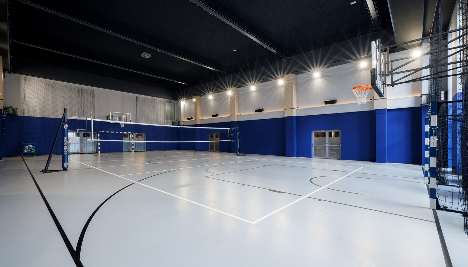 Indoor multi‑sport court with volleyball net, basketball hoops and handball goals; bright modern gym for guest recreation.