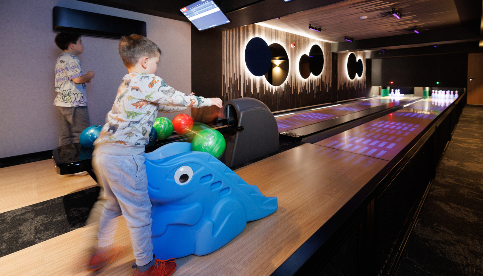 Hotel family bowling alley with two lanes, mood lighting, overhead score screens and a blue child ramp as kids roll colorful balls.