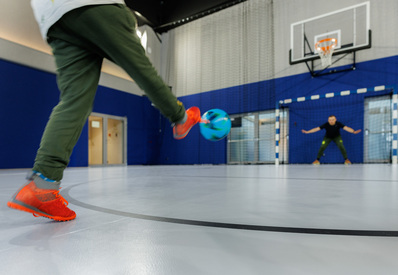 Vnitřní sportovní hala: host v oranžových teniskách kopne barevný míč směrem k brankáři pod basketbalovým košem.