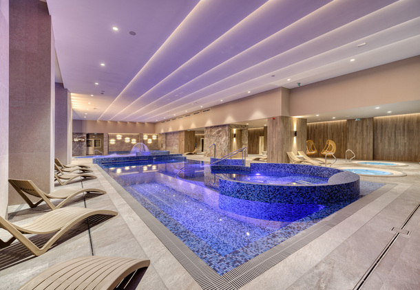 Luxusní vnitřní wellness s mozaikovým bazénem a vyvýšeným whirlpoolem, fialové osvětlení, dřevěné stěny a lehátka.