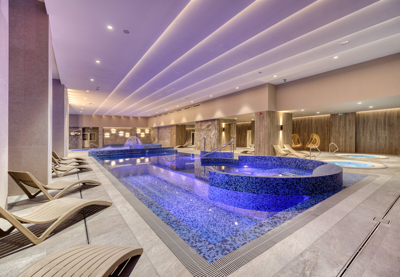 Luxusní vnitřní wellness s mozaikovým bazénem a vyvýšeným whirlpoolem, fialové osvětlení, dřevěné stěny a lehátka.