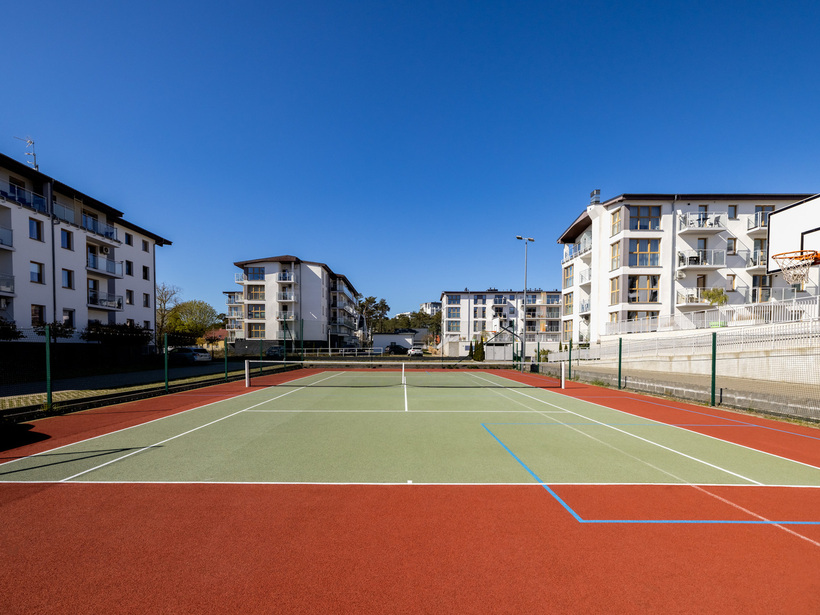 Venkovní multifunkční hřiště (tenis a basketbal) s červeno-zeleným povrchem, obklopené moderními budovami s balkony.