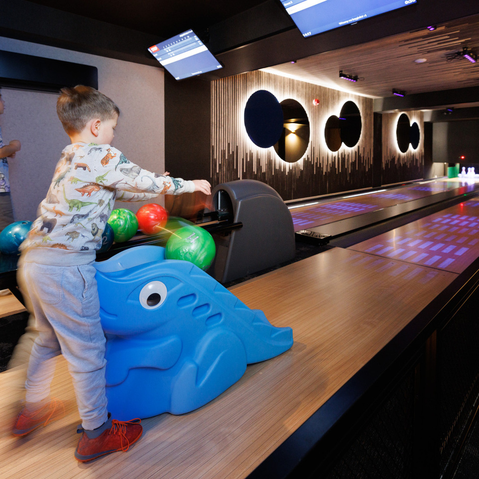 Rodinný bowling v hotelu se dvěma dráhami, náladovým osvětlením, obrazovkami skóre a modrou dětskou rampou, děti hází barevné koule.