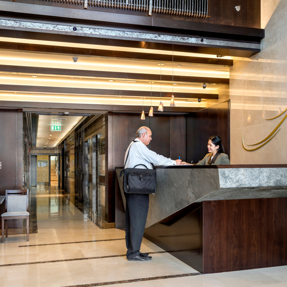 Modern hotel reception with dark wood and stone desk, warm lighting, staff assisting a guest checking in, and floral arrangement