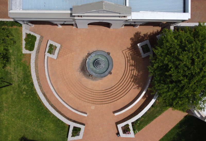 Aerial view of semicircular terracotta courtyard with tiered central fountain, curved steps, stone benches and leafy trees.