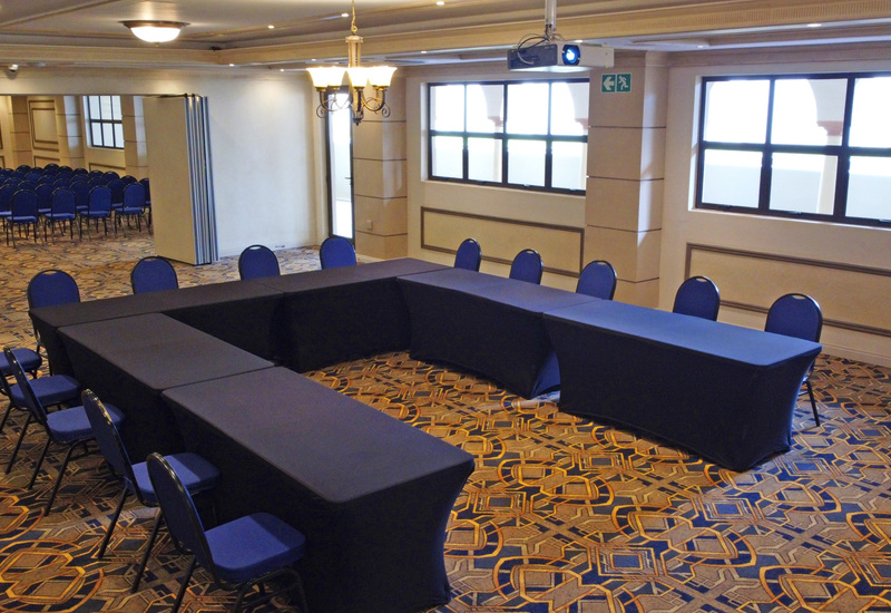 U-shaped meeting setup with navy table covers, blue chairs, ceiling projector and chandelier in a bright hotel function room