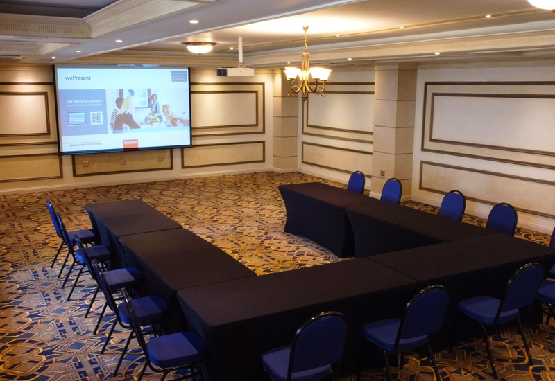 U-shaped meeting room with black-draped tables, blue chairs, ceiling projector and screen, patterned carpet and chandelier