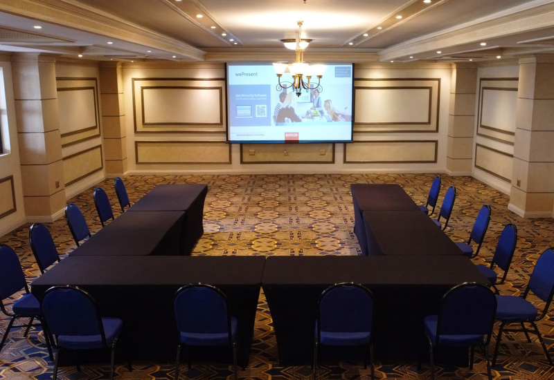 U-shaped meeting room with navy chairs, black-draped tables, chandelier, large projection screen and patterned carpet.